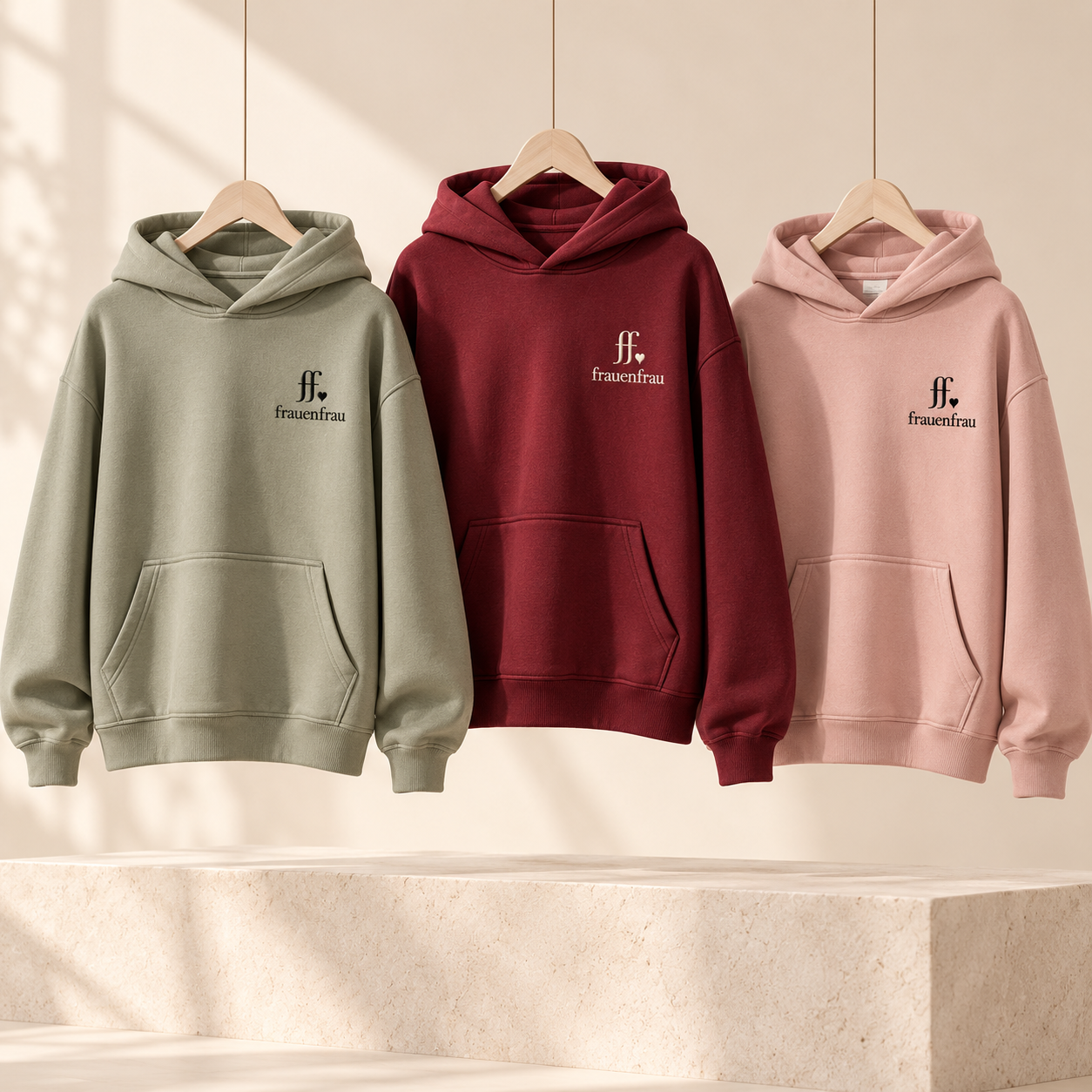 Drei hochwertige Hoodies in Salbeigrün, Bordeaux und Rosé mit ff-Herz-Logo und frauenfrau Schriftzug auf der linken Brust, auf Holzbügeln präsentiert